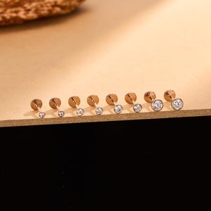 Könnte beinhalten: Eine Sammlung goldfarbener Ohrstecker mit flachen Rückseiten und klaren, runden Edelsteinen. Die Ohrringe sind in einer Reihe angeordnet und zeigen verschiedene Edelstein-Größen. Die Ohrstecker werden auf einer hellbraunen Oberfläche präsentiert.