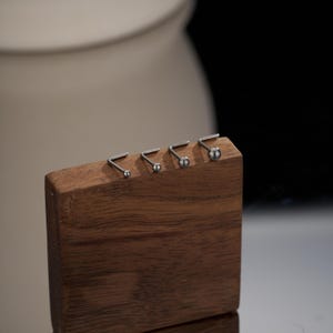 May include: Five silver nose rings of different sizes are displayed on a wooden stand. The rings are made of surgical steel and have a small ball at the end.