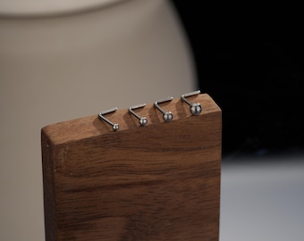 May include: Five silver nose rings of different sizes are displayed on a wooden stand. The rings are made of surgical steel and have a small ball at the end.