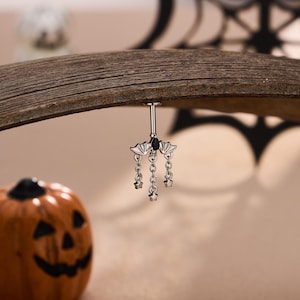 Könnte beinhalten: Ein silberfarbener Bauchnabelpiercing mit einem fledermausförmigen Anhänger, der in der Mitte einen kleinen schwarzen tropfenförmigen Stein trägt. Drei zarte silberne Ketten hängen an der Fledermaus, jede mit einem kleinen Sternanhänger.