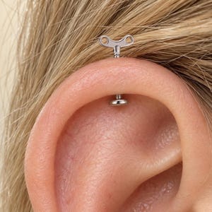May include: A silver-toned metal ear piercing stud featuring a wind-up key design. The jewelry consists of a flat circular back and a top piece shaped like a classic mechanical winding key with two circular openings, positioned in the ear cartilage.