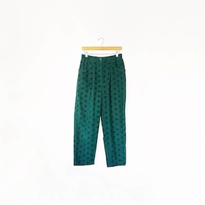 May include: Emerald green pants with a diamond pattern, hanging on a wooden hanger. The pants have a high waist and pockets. The fabric appears to be corduroy or a similar textured material. The background is plain white.