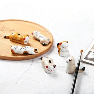 May include: A selection of ceramic cat-shaped chopstick rests in different poses. The cats are white, tan, and orange with black details. Some rests are inscribed with names such as "Aurora" and "Lily". A wooden tray and a paintbrush are also visible.