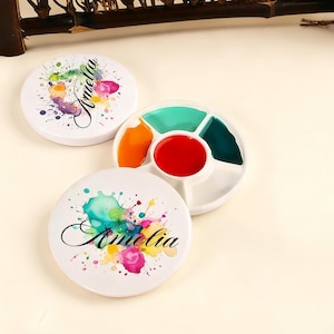 May include: White watercolor palette with a selection of vibrant colors, including red, orange, green, and blue. Two white round lids feature colorful paint splatter designs and the name "Amelia" in elegant script.