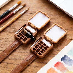 May include: Two brown watch-style paint palettes with open lids, each containing a small bottle and a brush. The watch bands are brown leather, one with "Isabella" and the other with "Margaret" engraved. Paintbrushes and a laptop are in the background.