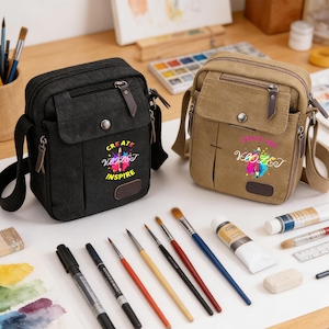 May include: Two canvas crossbody bags, one black and one tan, each featuring a front flap with a snap closure, a zippered pocket, and a graphic print that reads "CREATE INSPIRE" with a pencil and colourful paint splatters.