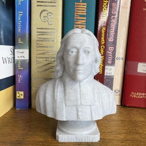 May include: A light grey bust of John Wesley, with his name inscribed on the base. The bust is positioned in front of a collection of books, including titles like "Faith & Holiness" and "The Doctrine of Sin."