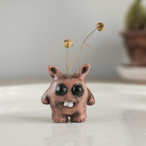 Op de afbeelding: Een klein, bruin, keramisch monsterfiguur met grote zwarte ogen en witte tanden. Het monster heeft twee gedroogde bloemstelen die uit zijn hoofd steken.