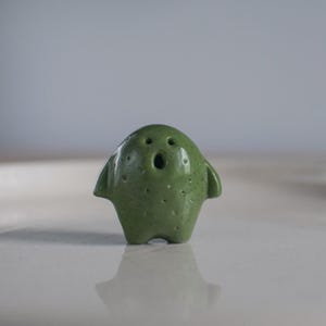 Könnte beinhalten: Eine kleine grüne Keramikfigur eines Wesens mit einem überraschten Ausdruck. Die Figur hat einen runden Körper und zwei kleine Arme.