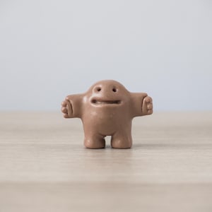 Peut inclure: Une petite figurine brun clair en forme de créature trapue et arrondie avec les bras tendus, deux petits yeux circulaires et une fente horizontale pour la bouche. La figurine a une texture lisse et mate et repose sur deux jambes courtes.