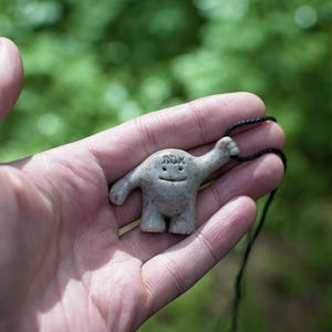 May include: A small, off-white ceramic pendant shaped like a cartoon figure with a smiling face and raised arm, strung on a black cord. The background is a blurred green.