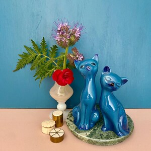Peut inclure: Deux figurines de chat en céramique bleu brillant sont posées sur une base verte imitation marbre. Un petit vase avec une fleur rouge et des fleurs violettes est à gauche, avec de petites boîtes décoratives.