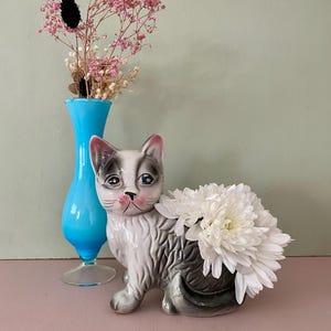 Peut inclure: Un vase en céramique en forme de chat gris et blanc, avec des accents roses sur les oreilles et le nez, rempli de fleurs blanches. Un vase turquoise avec des fleurs roses est derrière le vase chat. Le fond est un mur vert clair et une surface rose.