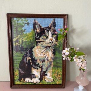 May include: Framed needlepoint art featuring a seated cat with black, white, and tan fur. The cat is set against a backdrop of a green field, trees, and a blue sky. The artwork is enclosed in a dark brown wooden frame.