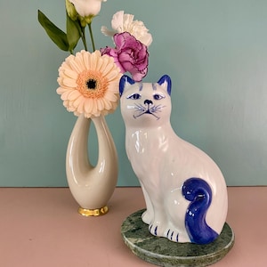 Op de afbeelding: Een witte keramische kattenfiguur met blauwe accenten, zittend op een groene marmeren voet. Een crèmekleurige vaas met gouden rand bevat een boeket bloemen, waaronder een perzikgerbera en witte en paarse bloemen.