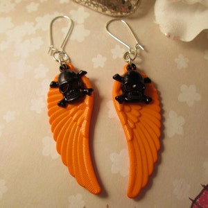 Könnte beinhalten: Ein Paar orangefarbene Engelsflügel-Ohrringe mit schwarzen Totenkopf-Anhängern. Die Ohrringe sind aus Kunststoff und haben silberne Haken.