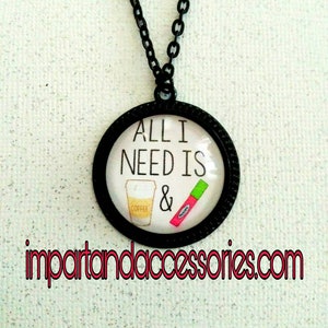 Op de afbeelding: Zwarte ketting met een ronde hanger met de tekst "All I need is &" met een afbeelding van een koffiebeker en lippenstift.