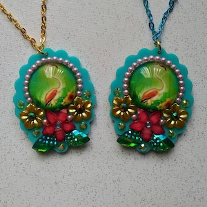 Peut inclure: Deux colliers pendentifs turquoise avec un motif de flamant rose. Chaque pendentif présente un cabochon rond en verre transparent avec un flamant rose dans un décor tropical. Les pendentifs sont entourés de feuilles dorées et vertes, de fleurs roses et rouges et de strass transparents.