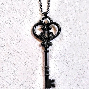 May include: Black key-shaped pendant necklace with a crown design and a clear rhinestone. The necklace has a black chain.