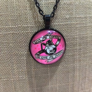 以下が含まれることがあります： 黒いチェーンネックレスにピンクのガラス製のペンダント。ペンダントには、骸骨と交差した骨を持つコーヒーポットの白黒のイラストが描かれています。ペンダントのテキストは「Give me coffee or give me death.」です。