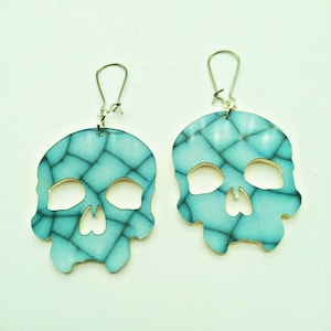 May include: A pair of skull-shaped earrings. Each earring is a light blue color with a cracked pattern design. The earrings have silver-colored ear wires.