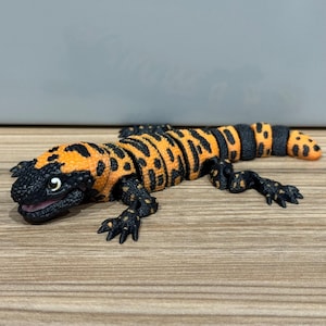 May include: A detailed figurine of a Gila monster, with a black head and limbs, and a body patterned with orange and black spots. The lizard is posed on a wooden surface.
