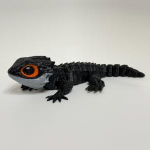 May include: A black and grey reptile figurine with a detailed, textured design. The figurine has large orange eyes and a segmented body, resembling a crocodile skink. The figure is posed on a white surface.