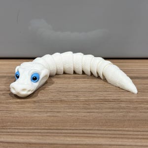 May include: A white segmented snake toy with blue eyes. The snake is curled up on a wooden surface.