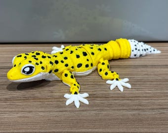 May include: A yellow and white articulated lizard figurine featuring black spots across its head and body. The creature has a white underside, white feet with distinct toes, and a segmented tail that transitions to a white tip with black spots.