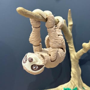 May include: A brown and beige sloth toy hanging upside down from a branch. The sloth has a textured fur pattern and large, brown eyes. The toy is made of plastic and is articulated at the joints.