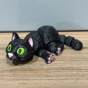 May include: A black cat figurine with green eyes and pink inner ears and paw pads. The cat is lying down on a wooden surface. The cat's tail is curved. The figurine is 3D printed.