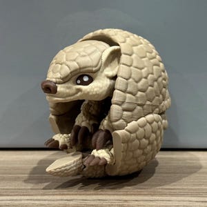 May include: A tan-coloured, 3D-printed pangolin figurine. The pangolin is curled up, with detailed scales and claws. It has a brown nose and dark eyes. The background is a neutral grey.