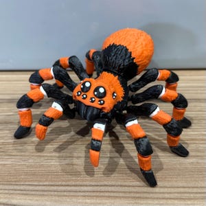 May include: A black and orange spider toy with large eyes and hairy legs. The spider is made of plastic and has a realistic appearance.
