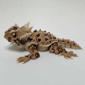 May include: A detailed 3D-printed model of a horned lizard, in tan and brown. The model has intricate details, including spikes, scales, and realistic eyes. The lizard is posed in a resting position, showcasing its legs and tail.