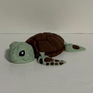 May include: A 3D-printed turtle figurine. The turtle has a brown shell, a light green head, and light green flippers with brown spots. The turtle is lying on a white surface.