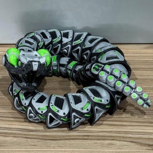 May include: A 3D-printed articulated snake toy with a futuristic design. The snake has a segmented body with grey, black, and green accents. The head and tail have glowing green orbs, giving it a sci-fi aesthetic.