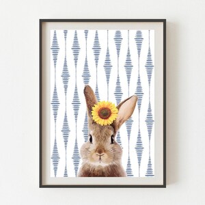 May include: Framed print of a brown rabbit with a yellow sunflower crown, set against a white and blue geometric pattern. The artwork is in a black frame. The print is suitable for a child's room.