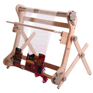 May include: A wooden tapestry loom with a colorful woven piece depicting buildings. The loom has adjustable parts and a light wood finish. The woven piece features red, orange, green, and blue hues.