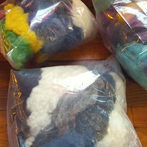 May include: Three clear plastic bags filled with assorted loose wool roving fibers in various colors including bright yellow, green, white, dark blue, and purple. The soft, fluffy material is packed into individual transparent storage pouches.