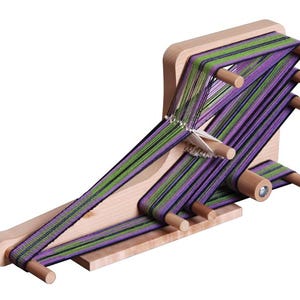 May include: A wooden weaving loom with green and purple yarn. The loom has a rectangular base and several wooden pegs to hold the yarn in place. The yarn is woven in a striped pattern.