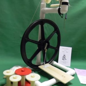 Pode incluir: Um torno de fiar de PVC branco com uma roda preta e um pedal de madeira. O torno de fiar tem uma bobina branca e um flyer branco. Há quatro bobinas brancas com tampas vermelhas na mesa abaixo do torno de fiar.