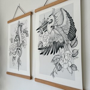 May include: Two framed art prints featuring detailed black and white illustrations. One print depicts a snake coiled among flowers, the other a bird with outstretched wings, also surrounded by flowers. The prints are held by wooden bars.