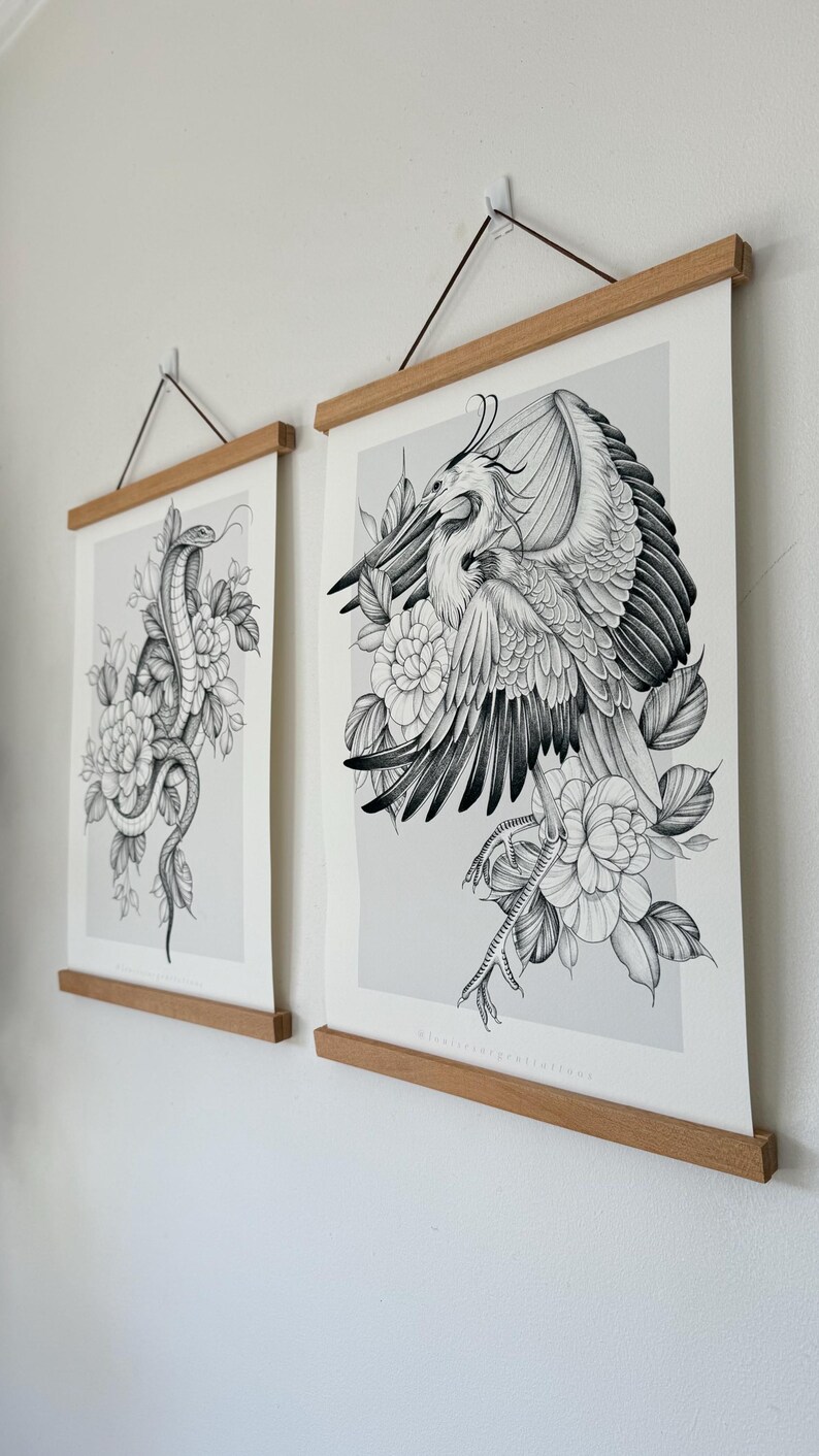 May include: Two framed art prints featuring detailed black and white illustrations. One print depicts a snake coiled among flowers, the other a bird with outstretched wings, also surrounded by flowers. The prints are held by wooden bars.
