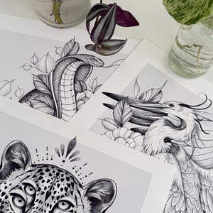 May include: Black and white art prints featuring detailed illustrations of a cobra, a leopard with multiple eyes, and a bird. The prints are displayed on a white surface, with plants in glass vases in the background. The art style is detailed and realistic.