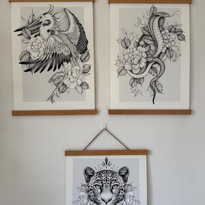 May include: Three framed art prints featuring detailed black and white illustrations. The top prints depict a heron and a cobra with floral accents. The bottom print showcases a leopard's face surrounded by flowers, all mounted on wooden hangers.