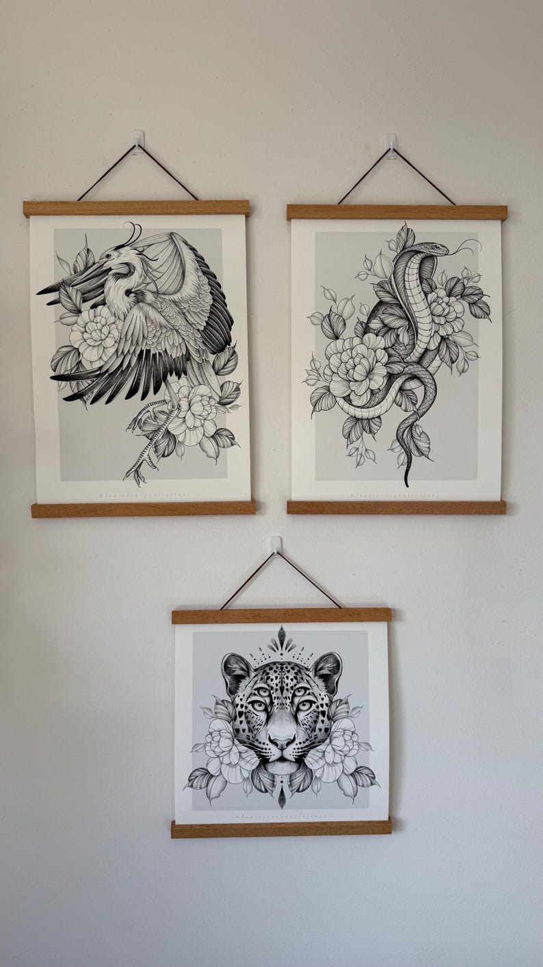 May include: Three framed art prints featuring detailed black and white illustrations. The top prints depict a heron and a cobra with floral accents. The bottom print showcases a leopard's face surrounded by flowers, all mounted on wooden hangers.