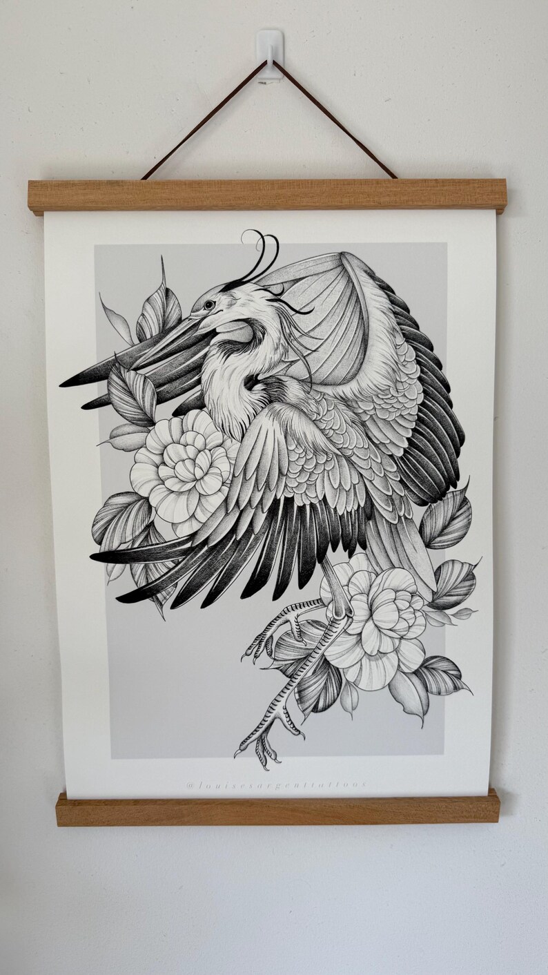 May include: Black and white illustration of a heron with outstretched wings, surrounded by flowers and leaves. The artwork is displayed on a white paper, held by a wooden frame. The artist's name is visible at the bottom.
