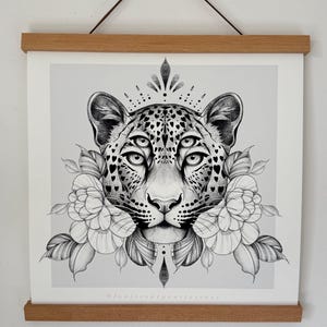 May include: Black and white illustration of a leopard with multiple eyes, adorned with floral elements and a crown-like design. The artwork is displayed on a light gray background and framed with wooden bars.