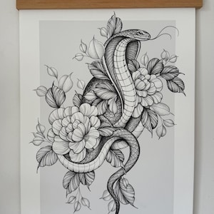 May include: Black and white illustration of a cobra coiled around flowers and leaves. The snake's tongue is extended. The artwork is displayed on a white paper with a wooden frame and hanging string.