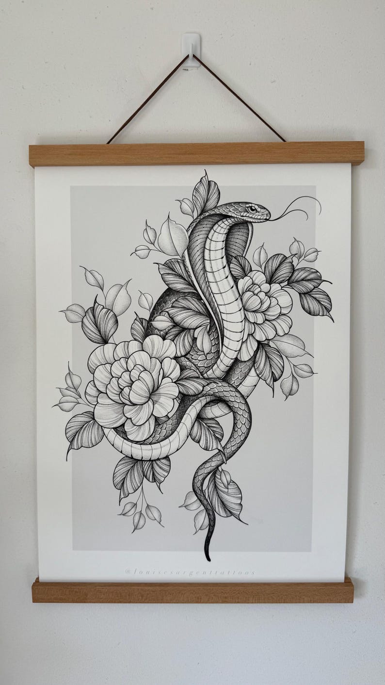 May include: Black and white illustration of a cobra coiled around flowers and leaves. The snake's tongue is extended. The artwork is displayed on a white paper with a wooden frame and hanging string.